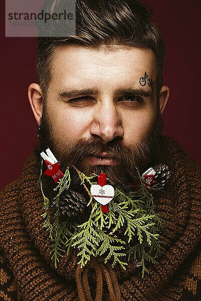 Lustiger bärtiger Mann in einem Neujahrsbild als Weihnachtsmann mit Dekorationen auf seinem Bart. Fest der Weihnacht. Fotos im Studio aufgenommen