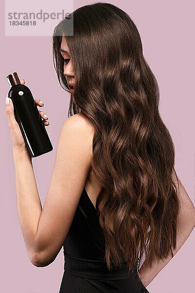 Schöne brünette Frau mit Locken und klassischem Make-up in einem schwarzen Kleid mit einer Flasche Shampoo in den Händen. Schönes Gesicht. Foto im Studio aufgenommen