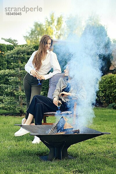 Zwei Freunde entspannen sich im Garten und unterhalten sich in der Nähe des Grills mit brennenden Holzscheiten (Fokus auf den Vordergrund)