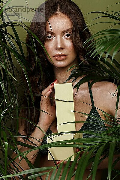 Schöne sexy Frau im Bikini posiert in der Mitte der grünen Pflanzen mit einer Box von Kosmetika in der Hand. Sommer-Look. Schönes Gesicht. Foto im Studio aufgenommen