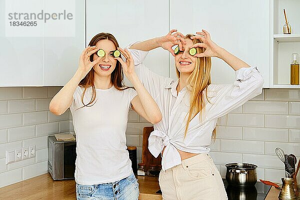 Zwei verspielte Frauen halten sich Gurkenstücke an die Augen und haben Spaß in der Küche