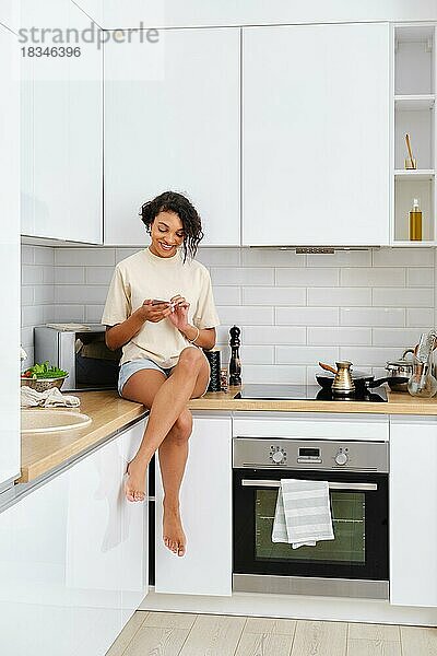Junge Frau sitzt am Küchentisch  liest auf dem Handy und wartet darauf  dass der Kaffee auf dem Herd gebrüht wird