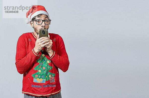 Mann in Weihnachtsmütze mit erstauntes Gesicht hält Telefon. Online Weihnachten Sonderangebote Konzept. Junger Mann in weihnachtlicher Kleidung und Hut erstaunt hält das Telefon