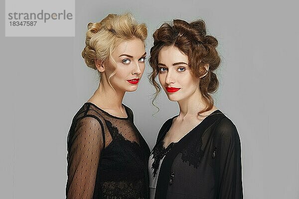 Zwei hübsche Mädchen mit lockigem Haar flirten. Blonde und brünette Mädchen mit natürlichem Make-up und roten Lippen