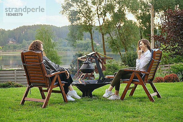 Zwei Frauen entspannen sich im Garten  sitzen auf Stühlen und unterhalten sich in der Nähe des Grills