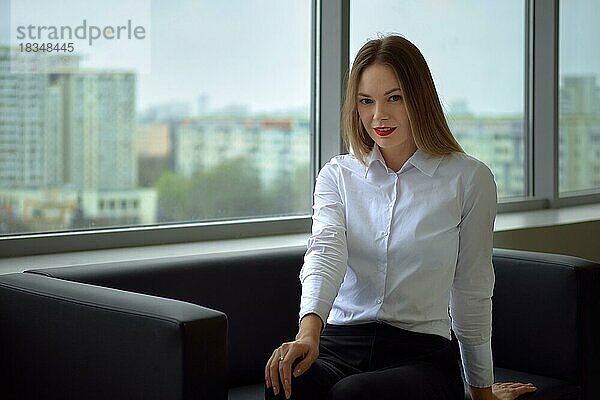 Schöne und positive Geschäftsfrau auf der Couch schaut gerade und lächelnd  Fenster mit Stadtlandschaft im Hintergrund