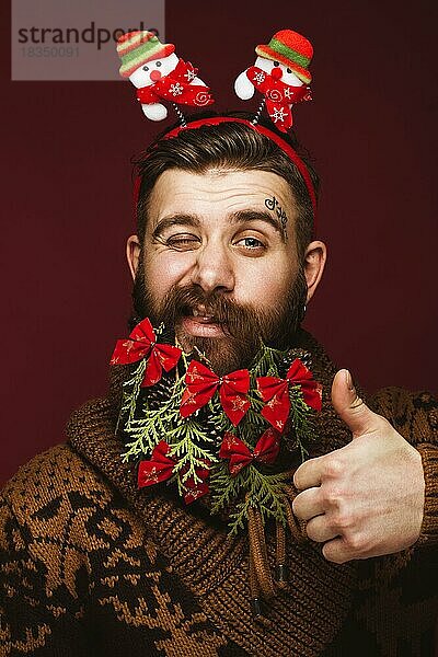 Lustiger bärtiger Mann in einem Neujahrsbild als Weihnachtsmann mit Dekorationen auf seinem Bart. Fest der Weihnacht. Fotos im Studio aufgenommen