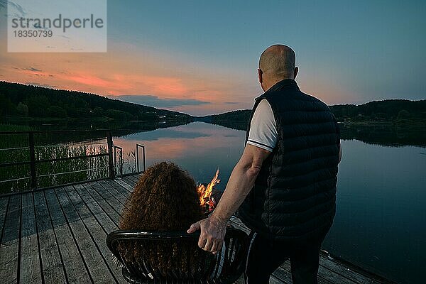 Niedliches Paar beobachtet den Sonnenuntergang am Feuer auf dem Pier (Blick von hinten)