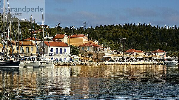 Morgenlicht  Hafenpromenade  Segelboote  Häuser  grüne Hügel  blauer Himmel mit grauweißen Wolken  Hafenort  Fiskardo  Insel Kefalonia  Ionische Inseln  Griechenland  Europa