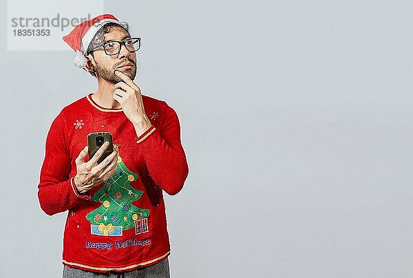 Hübscher Mann in Weihnachtskleidung nachdenklich hält Handy. Nachdenklicher Mann mit Weihnachtsmütze hält Handy  Junger Mann in weihnachtlicher Kleidung nachdenklich mit Handy isoliert