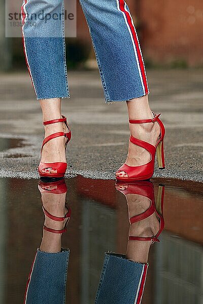 Reflexion von Frauenbeinen in Jeans und roten Schuhen in einer Pfütze auf Asphalt