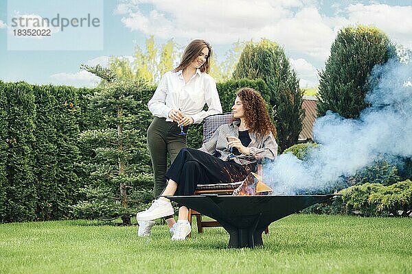 Zwei Freunde entspannen sich im Garten und unterhalten sich in der Nähe des Grills mit brennenden Holzscheiten