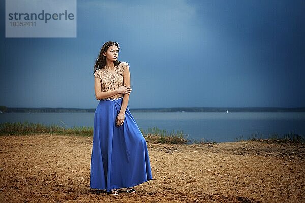 Ruhige einsame Mode-Modell in langen blauen Rock und Spitze Bluse stehend auf einem Strand in einem bewölkten Tag