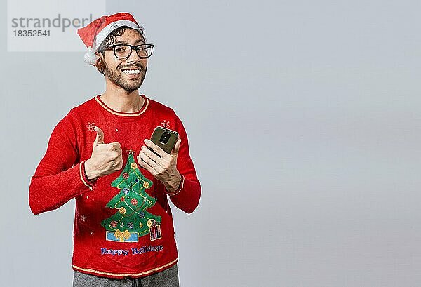 Lächelnder Mann in Weihnachtsmütze mit Handy und Daumen hoch. Lächelnder junger Mann in Weihnachtsmütze mit Handy und geben Daumen nach oben. Menschen in santa Hut mit Telefon und tun ok Geste