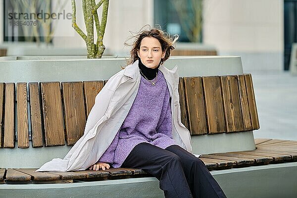 Junge Frau in weiten Hosen  Pullover  Regenmantel und groben Stiefeln sitzt auf einer Bank und wartet auf einen Freund