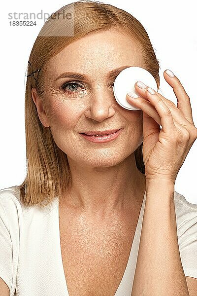 Porträt einer schönen älteren Frau in einem weißen Hemd  die sich mit einem Reinigungsmittel und einem Wattepad abschminken will