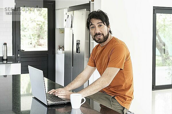 Lateinamerikanischer Mann  der in der Küche an seinem Laptop arbeitet