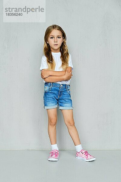 Kleines Mädchen in weißem T-Shirt  Jeans-Shorts und rosa Turnschuhen steht mit gekreuzten Händen auf der Brust in der Nähe der Wand