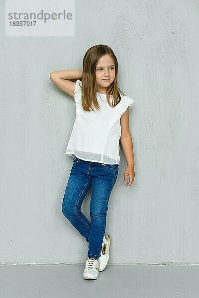 Kleines Mädchen in weißer Bluse und Jeans lehnt sich an die Wand und hält eine Hand hinter den Kopf