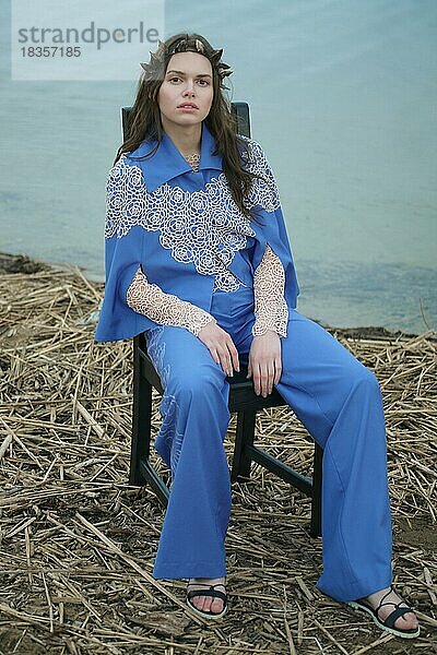 Ruhige einsame Mode-Modell in blauen Hosen und Umhang sitzen auf einem Stuhl in einem bewölkten Tag am Strand
