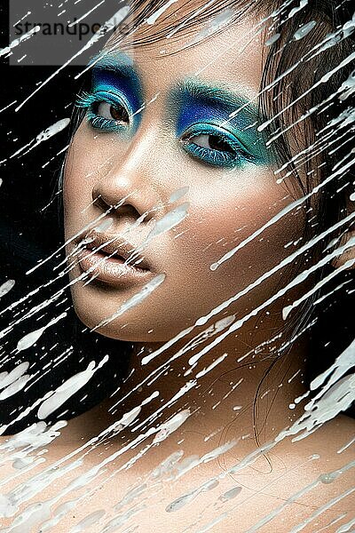Schönes asiatisches Mädchen mit hellblauem Make-up hinter Glas mit Wachstropfen. Schönes Gesicht. Bild im Studio auf einem schwarzen Hintergrund genommen