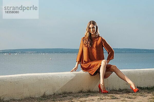 Hübsches Mädchen mit langen schlanken Beinen in Seidenkleid sitzt auf Pier mit Meer auf Hintergrund