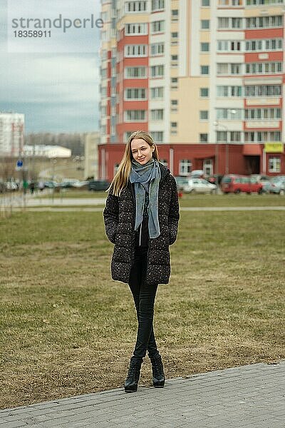 Fröhliches Mädchen in Jeans und Daunenjacke geht die Straße in einem Wohngebiet mit hohen Häusern entlang