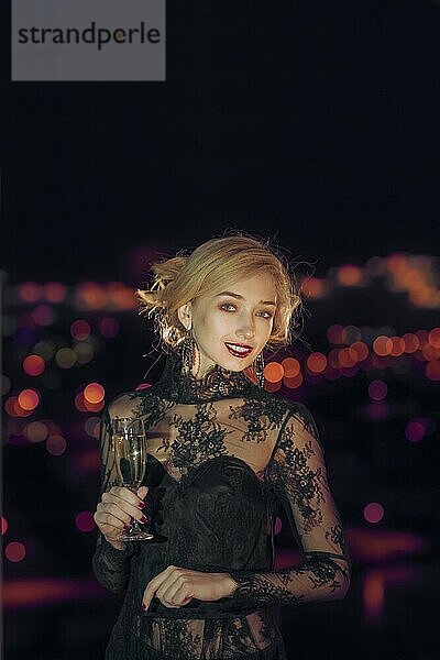 Schönes Mädchen mit Glas Champagner stehen in der Nähe des Fensters mit unscharfen Nacht Stadt auf Hintergrund