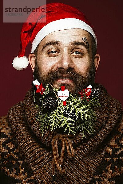 Lustiger bärtiger Mann in einem Neujahrsbild als Weihnachtsmann mit Dekorationen auf seinem Bart. Fest der Weihnacht. Fotos im Studio aufgenommen