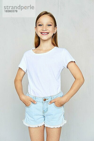 Junges sportliches Mädchen in weißem T-Shirt mit Händen in Jeans-Shorts posiert im Studio