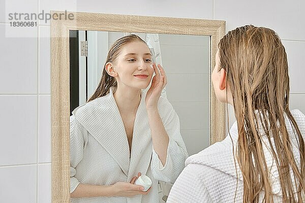 Junge Frau steht vor dem Spiegel und trägt nach dem Duschen Feuchtigkeitscreme auf das Gesicht auf. Hautpflege tägliche Routine