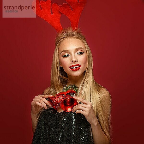 Schönes blondes Mädchen in einem Neujahrsbild mit Weihnachtsglocken um ihren Hals und Hirschhörnern auf ihrem Kopf. Schönes Gesicht mit festlichem Make-up. Foto im Studio aufgenommen