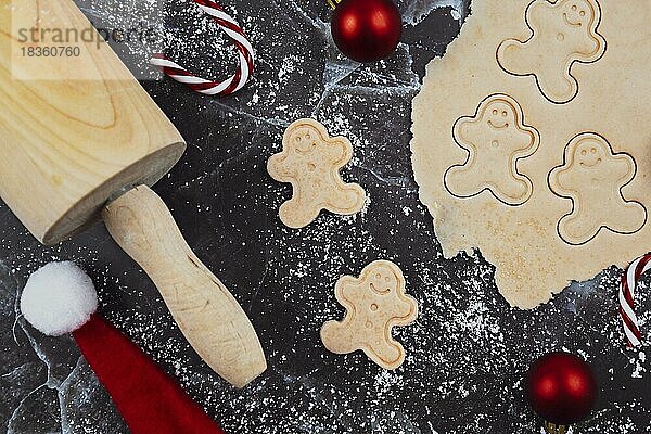 Flat Lay Konzept für das Backen von Weihnachtsplätzchen mit ausgerollten Plätzchenteig  Kekse in Form von glücklichen Lebkuchenmänner und saisonale Dekoration wie Kugeln auf dunklem Marmor Hintergrund
