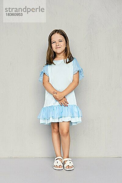 Nettes kleines Mädchen in blauem Kleid und weißen Sandalen posiert mit den Händen verschränkt unten in der Nähe der grauen Wand