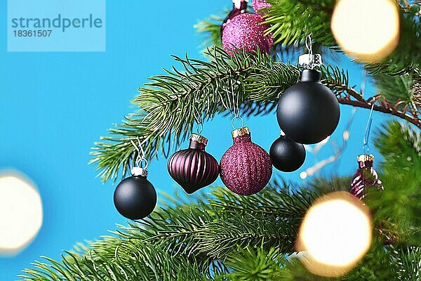 Close up von lila und schwarz Glas Baumkugel mit geschmückten Weihnachtsbaum mit anderen saisonalen Baumschmuck auf blauem Hintergrund und Weihnachten Licht Bokeh im Vordergrund