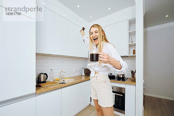 Fröhliche Frau tanzt und singt mit einer Tasse Kaffee in der Hand in der Küche