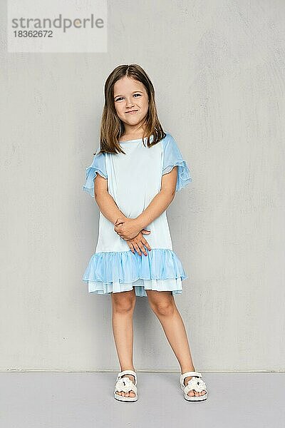 Nettes kleines Mädchen in blauem Kleid und weißen Sandalen posiert mit den Händen verschränkt unten in der Nähe der grauen Wand