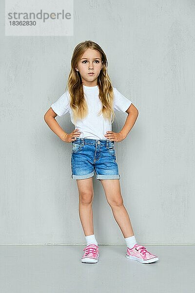 Kleines Mädchen in weißem T-Shirt  Jeans-Shorts und rosa Turnschuhen steht mit den Händen auf der Taille in der Nähe der Wand