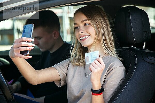 Glücklich lächelndes Mädchen macht ein Selfie mit einem neuen Führerschein  neben einem Fahrlehrer sitzend