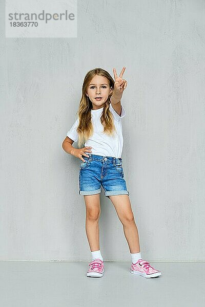 Kleines Mädchen in weißem T-Shirt  Jeans-Shorts und rosa Turnschuhen stehend zeigt V-Zeichen