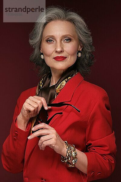 Porträt einer schönen älteren Frau in einer roten Jacke mit Accessoires und klassischem Make-up und grauem Haar