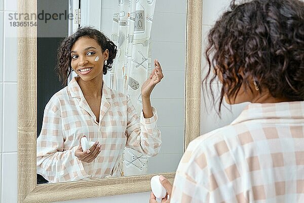 Lächelnde afroamerikanische Frau schaut in den Spiegel und trägt Gesichtscreme im Badezimmer auf