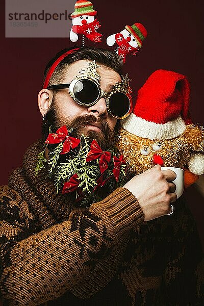 Lustiger bärtiger Mann in einem Neujahrsbild als Weihnachtsmann mit Dekorationen auf seinem Bart. Fest der Weihnacht. Fotos im Studio aufgenommen