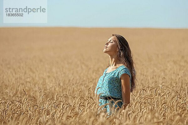Unbeschwertes Mädchen im blauen Sommerkleid steht unter der Sonne in einem Weizenfeld