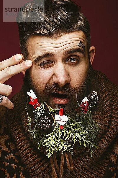 Lustiger bärtiger Mann in einem Neujahrsbild als Weihnachtsmann mit Dekorationen auf seinem Bart. Fest der Weihnacht. Fotos im Studio aufgenommen