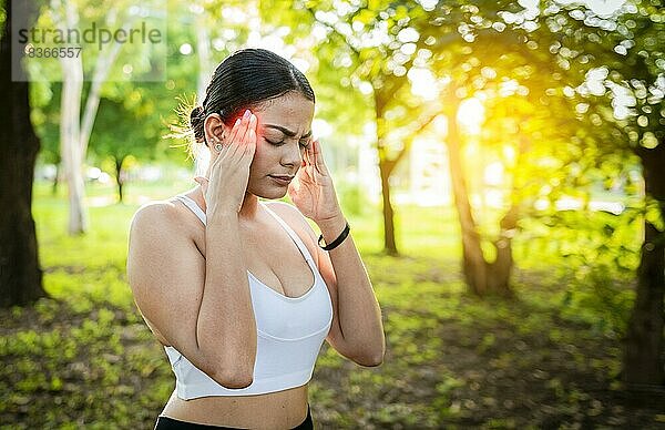 Junge Frau Läuferin reibt ihren Kopf mit Migräne in einem Park. Läuferin mit Kopfschmerzen und Müdigkeit im Park  Läuferin mit Kopfschmerzen im Park  Sportler Mädchen mit Migräne in einem Park