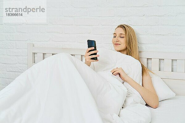 Junge  fröhliche Frau wacht auf  nimmt ihr Smartphone und liest den Feed der sozialen Medien