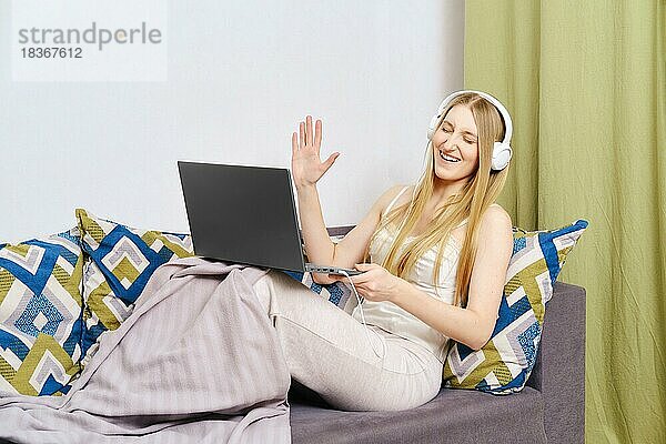 Positive Frau auf der Couch  die mit der Hand winkt  während sie mit Laptop und Kopfhörern zu Hause während einer Pandemie einen Videoanruf führt