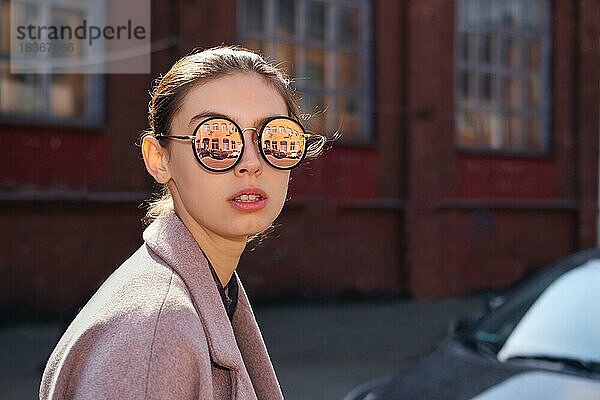 Junges Mädchen in offenem Mantel genießt die Sonne. Spiegel Sonnenbrille mit Straße reflektiert in ihm