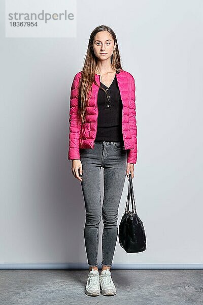 Ganzkörperporträt eines hübschen Mädchens in Daunenjacke und Jeans  das mit einer Handtasche an der Wand steht